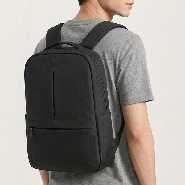 A man wearing a gray t-shirt and dark pants, shown from a side-back angle while wearing a black minimalist backpack against a light off-white background.