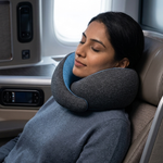 A woman rests in an airplane seat with her head supported by a gray U-shaped travel neck pillow with a blue inner lining.