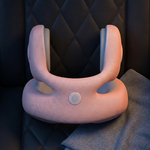 A light pink contoured neck pillow with a central circular control button, resting on a dark quilted leather car seat next to a folded gray blanket.