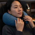 A woman wearing a dark gray knit sweater sits in an airplane seat, adjusting a blue textured travel neck pillow.