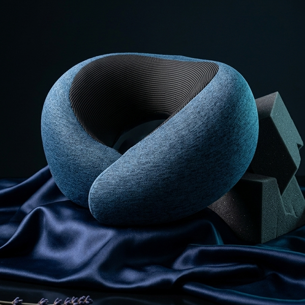 A blue-gray contoured neck pillow with a textured fabric exterior and ribbed dark interior, styled on draped navy satin fabric with lavender sprigs in the foreground.