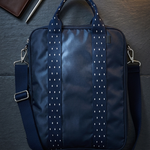 A navy blue nylon laptop bag with white dotted vertical stripes on the handles, styled flat-lay on a dark slate surface with a brown leather wallet, pen, and a lit candle in the background.