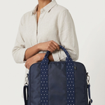 A woman wearing a light beige linen shirt and dark pants holds a navy blue tote bag with white polka-dot trim and handles against a plain off-white background.