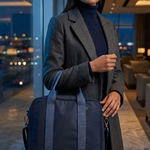 A woman in a gray coat and navy turtleneck holds a dark navy blue travel bag with light blue-gray striped handles in a modern, softly lit indoor setting with a city view at night.