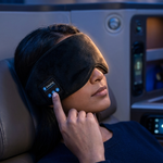 A woman reclining in a plane seat wearing a black sleep mask with a small digital control panel on the side, touching the strap to demonstrate its features.