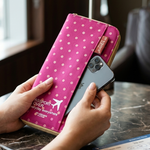 A person holds a pink passport cover with white polka dots and a gold border, sliding a smartphone into the slot. The cover has white text reading 'PASSPORT COVER' and a small airplane graphic.