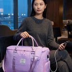 A woman sits in a modern lounge setting holding a lavender purple travel duffel bag with a matching shoulder strap. The bag has multiple zippered compartments and a small front label.