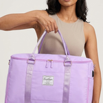 A woman holding a lavender purple duffel bag with dual top handles and a front zipper pocket. The bag has a small white rectangular brand patch on the front that reads 'Sofia'.