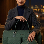 A woman in a dark gray blazer and navy turtleneck holds a dark green quilted duffel bag and a matching zip pouch on a dark marble table, with a softly blurred city window background.