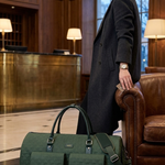 A woman in a dark coat stands in a hotel lobby next to a dark green textured duffel bag and a matching small pouch.