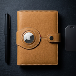 A tan leather planner or notebook cover with a circular metal emblem and snap closure, shown on a dark textured background alongside a black pen and a black rectangular accessory.