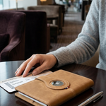 A tan leather travel document holder with a magnetic closure and a circular cutout, resting on a dark wooden table in a modern lounge setting. A person's hand rests on the holder next to a boarding pass, with a silver pen nearby.