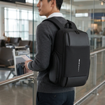 A man in a gray sweater and dark pants stands in an airport terminal wearing a black backpack, shown from the back. The backpack has a clean, minimalist design with a small vertical white logo on the front pocket.