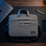 A gray fabric laptop bag with dual top handles and a shoulder strap, styled on a dark desk with a laptop, pen, and tablet nearby.
