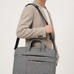 A man wearing a beige blazer and trousers carries a gray textured briefcase-style shoulder bag with a black strap against a plain light background.