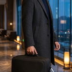 A man in a dark gray overcoat and navy turtleneck holds a black rectangular briefcase or laptop bag in a modern hotel lobby with large windows and warm floor lamps.