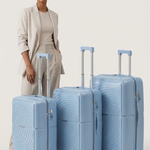 A woman in a beige blazer and matching pants stands beside three hard-shell spinner suitcases in a light dusty blue color, arranged by size against a plain light gray studio background.