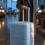 A woman in a gray coat stands in an airport terminal holding the handle of a light blue hard-shell spinner suitcase with a wavy circular pattern on the front.