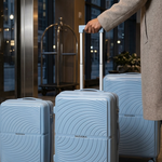 Three light blue hard-shell suitcases with a wavy circular pattern and spinner wheels are shown in a hotel lobby. A person wearing a beige coat is standing on the right, holding the handle of the middle suitcase.