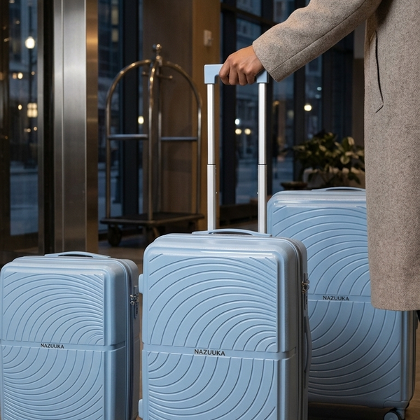 Three light blue hard-shell suitcases with a wavy circular pattern and spinner wheels are shown in a hotel lobby. A person wearing a beige coat is standing on the right, holding the handle of the middle suitcase.
