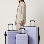 A woman in a beige blazer stands behind three hard-shell spinner suitcases in a matching light lavender color, set against a plain light gray studio background.