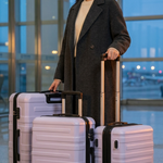 A woman in a dark coat stands in an airport terminal holding three light lavender hard-shell spinner suitcases with black trim.