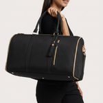 A woman in a black outfit carries a large black duffel bag with gold-tone zippers and handles against a light gray studio background.