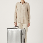 A woman stands next to a silver hard-shell spinner suitcase against a plain light gray studio background. She is wearing a light beige button-up shirt and matching beige pants.