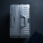 A top-down view of a metallic silver hard-shell suitcase on a dark textured surface, with a folded dark navy garment placed in the lower right corner.