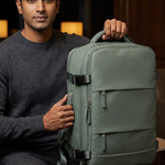 A man in a dark gray sweater sits holding a sage green laptop backpack in a dimly lit indoor setting.