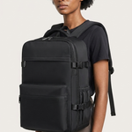 A woman with short dark curly hair wearing a black t-shirt and brown pants, carrying a large black backpack on her back against a light gray studio background.