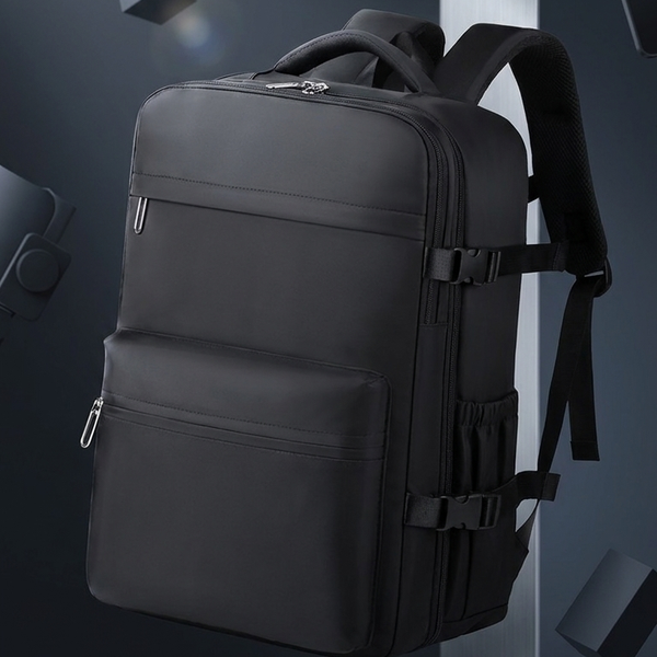 A black multi-compartment backpack with padded shoulder straps and side compression buckles, shown floating in a dark gray studio setting with geometric props.