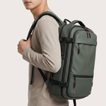 A man wearing a light beige long-sleeve shirt and dark pants is shown from the side, carrying a dark green backpack with black straps and red zipper pulls against a light gray studio background.