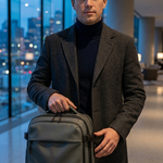 A man in a dark gray overcoat and black turtleneck holds a dark gray backpack with black straps in a modern indoor hallway with large windows overlooking a city at dusk.