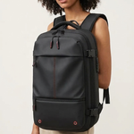 A woman with curly black hair wearing a black backpack with beige pants, photographed against a plain off-white studio background.