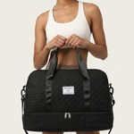 A woman with an afro hairstyle holds a black quilted duffel bag with a white brand label. She is wearing a white sports bra and shorts against a solid light beige background.