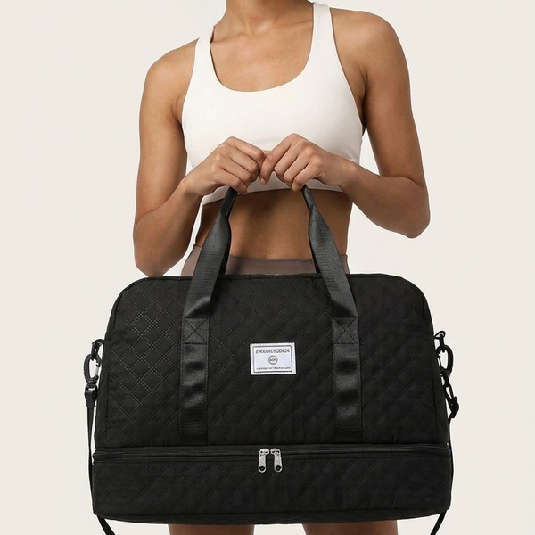 A woman with an afro hairstyle holds a black quilted duffel bag with a white brand label. She is wearing a white sports bra and shorts against a solid light beige background.