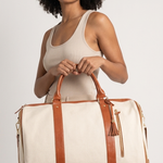 A woman holding a large cream canvas duffel bag with brown leather handles and straps, set against a plain light gray studio background.