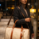 A woman in a dark gray coat carries a cream canvas duffel bag with brown leather trim and handles in a warmly lit hotel lobby.