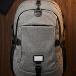 A gray heathered canvas backpack with black straps and trim, laid flat on a dark wood background with a tablet and wallet nearby.