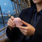 A woman in a dark navy blazer and shirt holds a small pink wireless earbud case while seated by large windows in a modern indoor space.