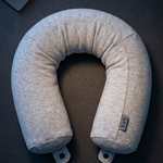 A light gray U-shaped travel neck pillow with a small fabric tag, styled on a dark textured surface with a passport and earbuds nearby.