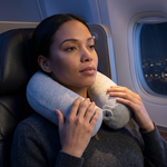 A woman sitting on an airplane seat using a light gray inflatable neck pillow for support, with the airplane window and cabin interior visible in the background.