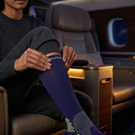 A woman seated in an airplane cabin puts on a dark blue crew sock with a geometric pattern and orange logo. The scene is lit by the warm interior cabin lights.