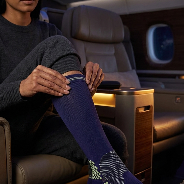 A woman seated in an airplane cabin puts on a dark blue crew sock with a geometric pattern and orange logo. The scene is lit by the warm interior cabin lights.