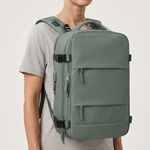 A woman wearing a light gray t-shirt and dark pants models a sage green backpack with multiple zippered compartments and top handles against a plain light gray background.