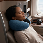 A woman rests her head on a blue travel neck pillow in a plush airplane business class seat, with an airplane window and tray table visible in the background.