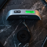 A digital pocket scale with a black strap and silver metal hook, resting on a dark marble surface with a pen, wallet, and phone nearby.