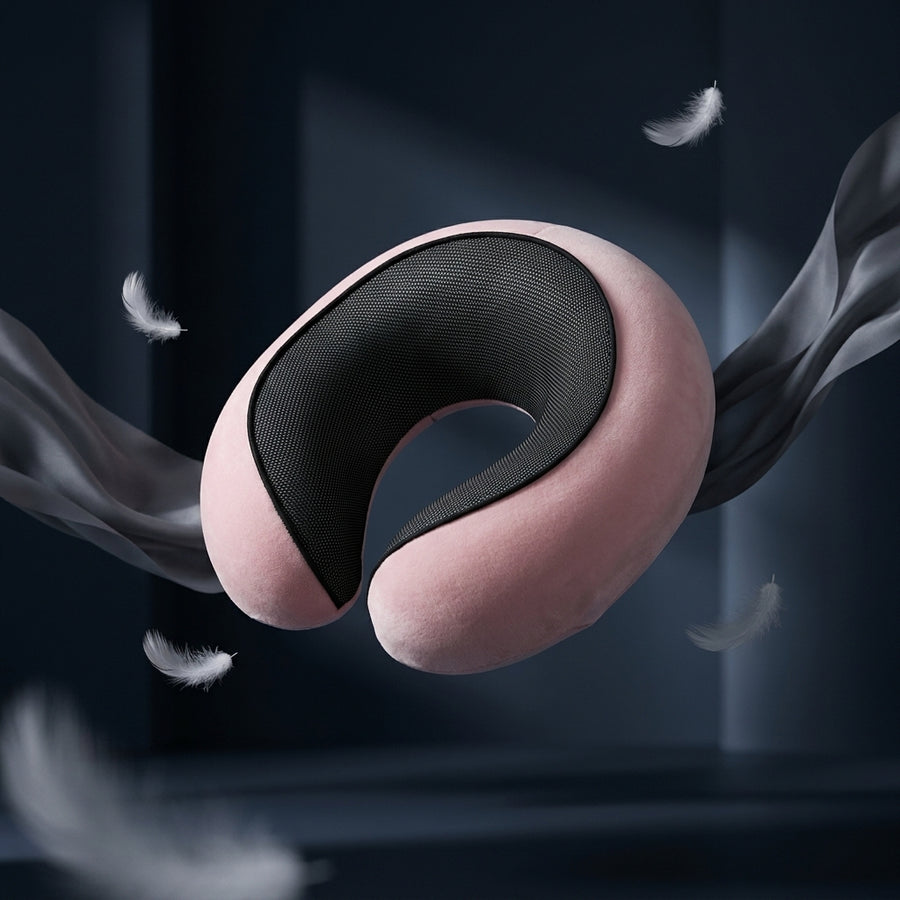 A U-shaped neck pillow with a soft pink exterior and a black textured inner cushion, shown floating against a dark blue-gray background with decorative feathers and flowing fabric around it.