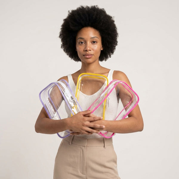 A woman with an afro hairstyle holds three clear plastic zippered pouches with purple, yellow, and pink borders against a plain light gray background.
